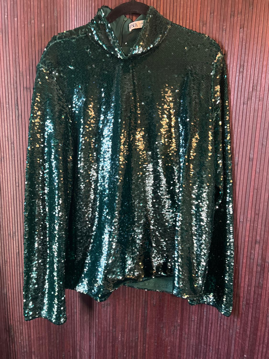 Green sequin blouse hanging on a hanger against a brown wooden background. Visible tag on the collar.