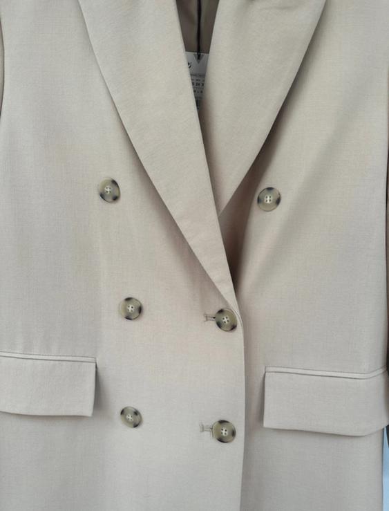 Beige double-breasted coat with wide lapels and front pockets. Visible label inside the collar.