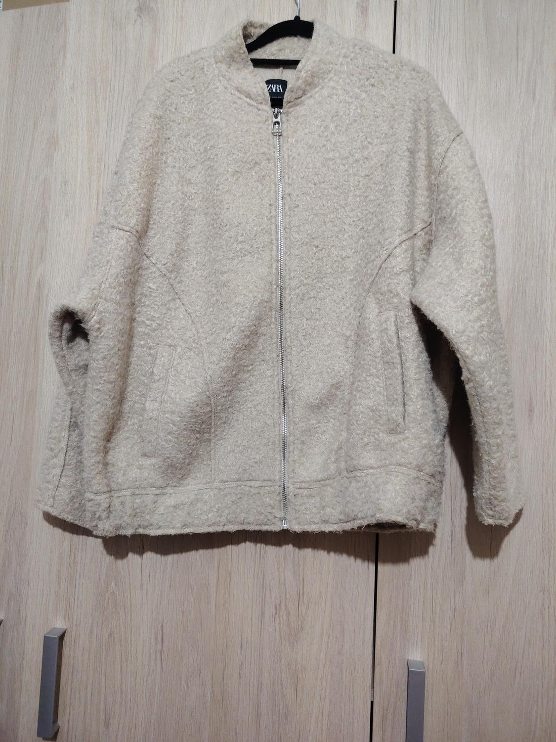 Soft-textured beige jacket with a front zip, hanging on a hanger against a light wooden background.