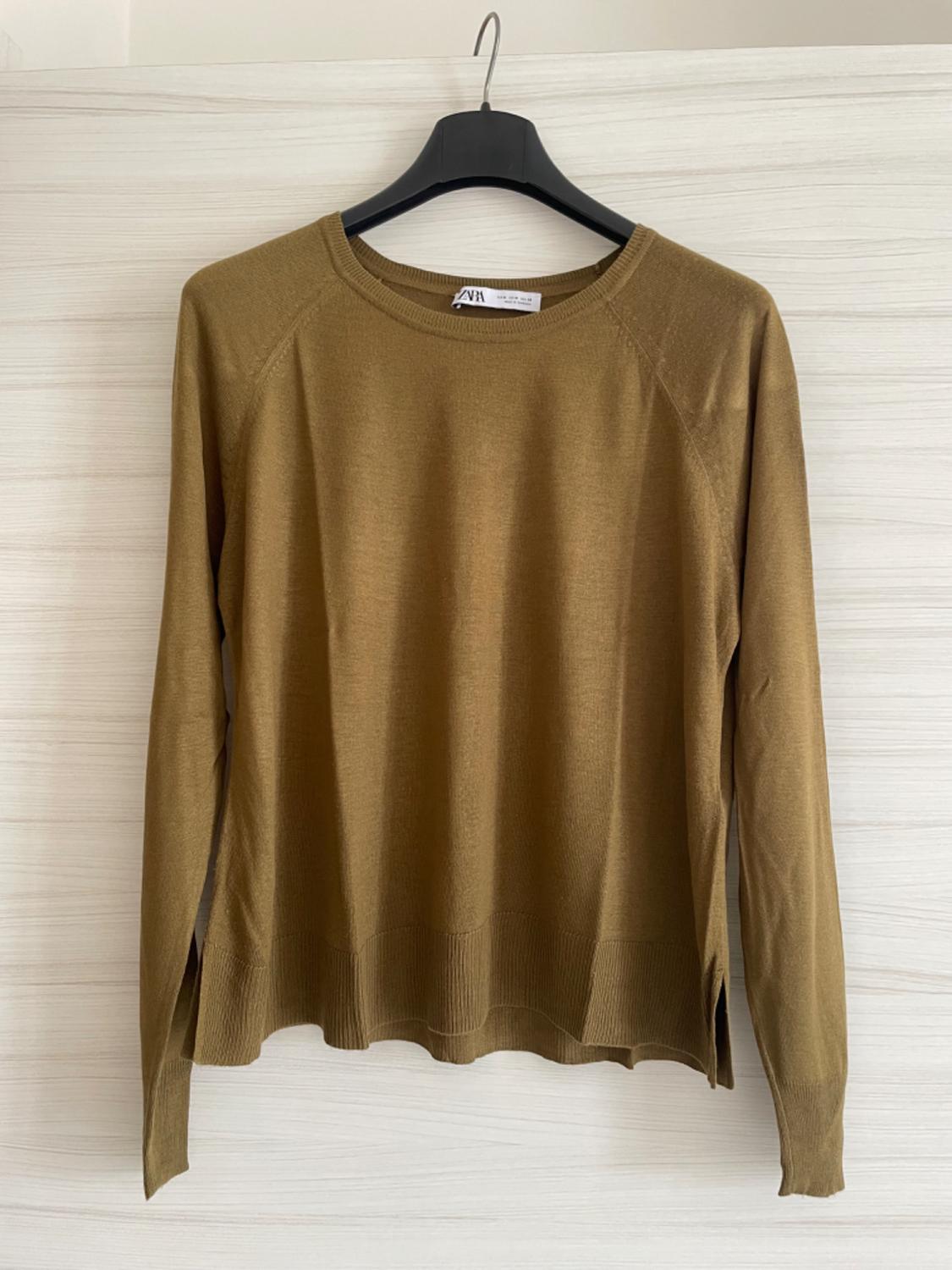 Brown long-sleeved jumper hanging on a black hanger against a light wooden background. Label visible on the collar.