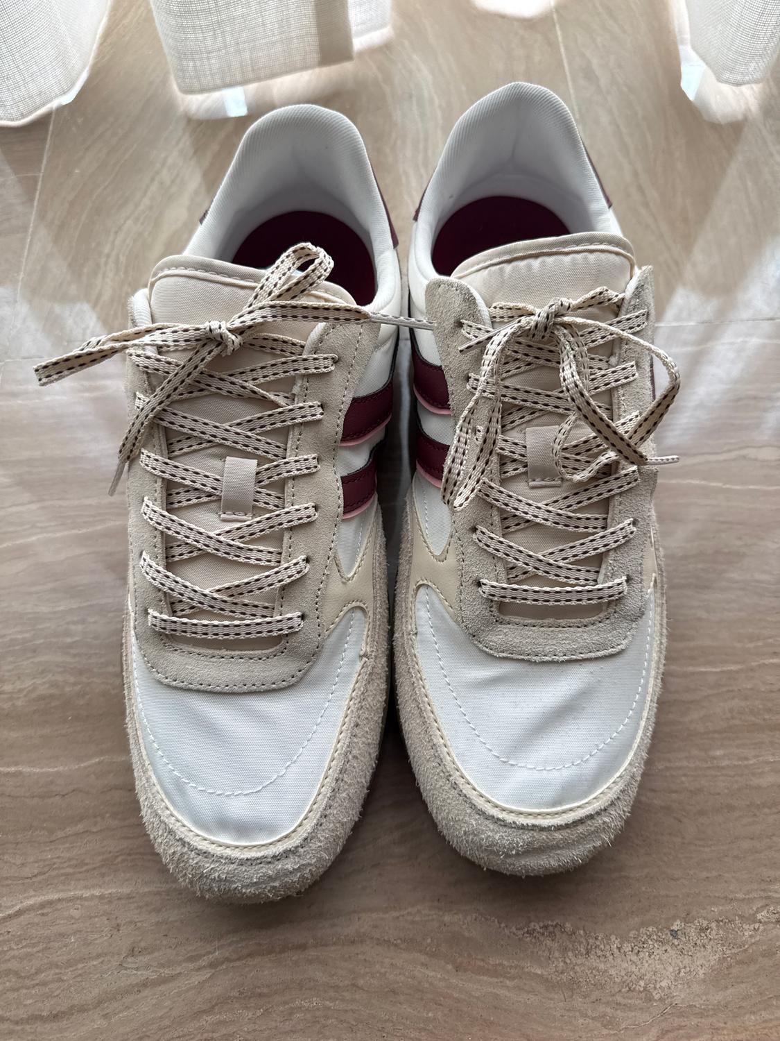 White sports trainers with beige and burgundy details, viewed from above on a light wooden floor. Beige laces with pattern.