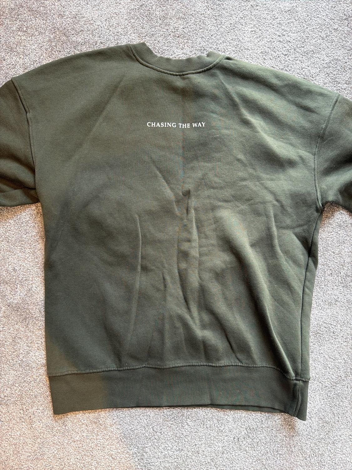 Dark green sweatshirt seen from the back on a grey carpet. It has the text "CHASING THE WAY" in white on the back.