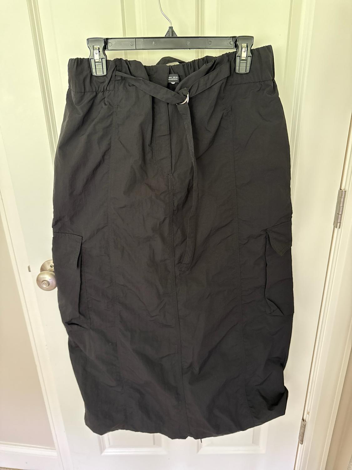 Black long cargo-style skirt with side pockets and a belt, hanging on a hanger in front of a white door.