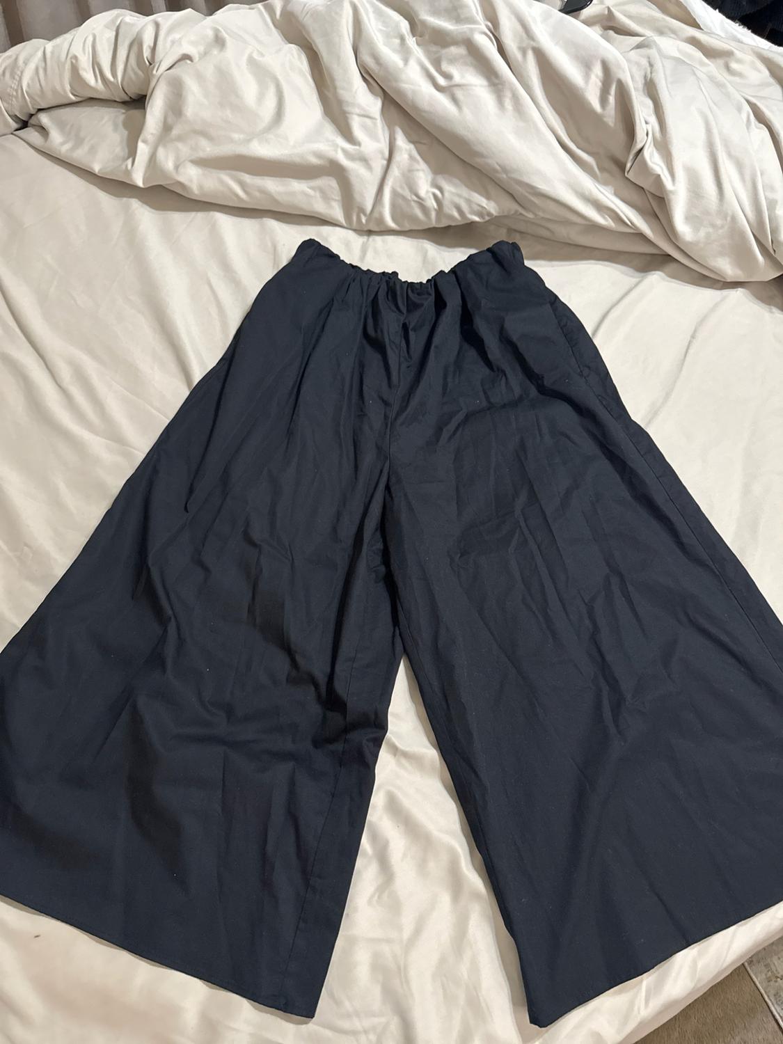 Very wide dark trousers with a gathered waist, laid out on a bed with light-coloured sheets. Casual style.