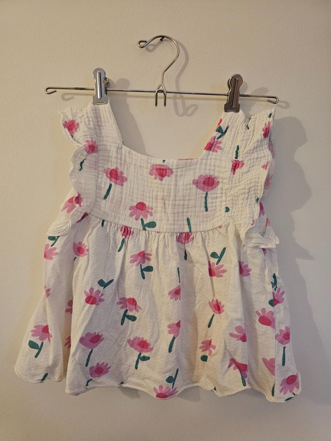 White blouse with pink and green floral print, hanging on a metal hanger against a beige wall.