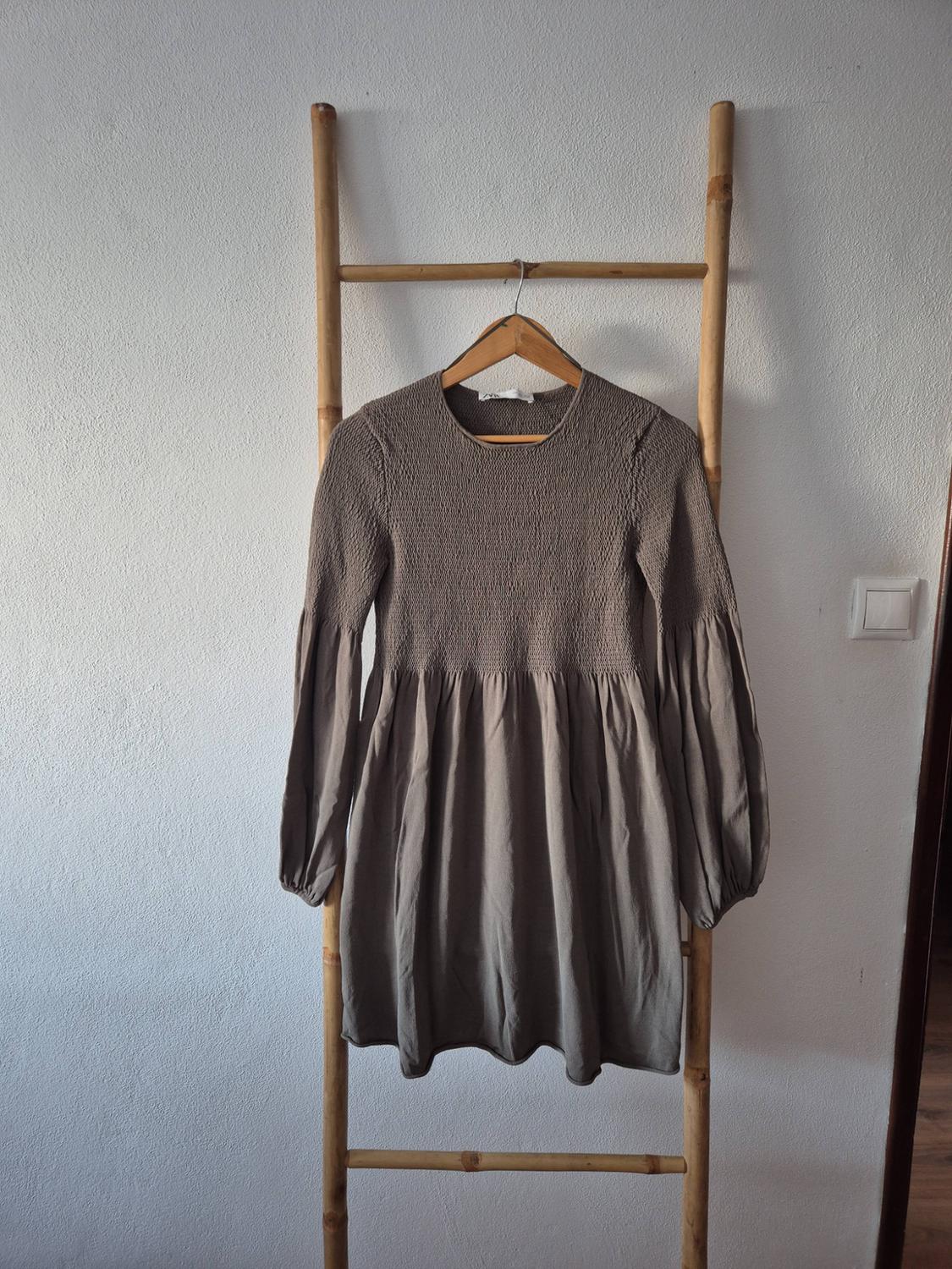 Short brown dress with long puffed sleeves and a ruched bodice, hanging on a wooden ladder leaning against a white wall.