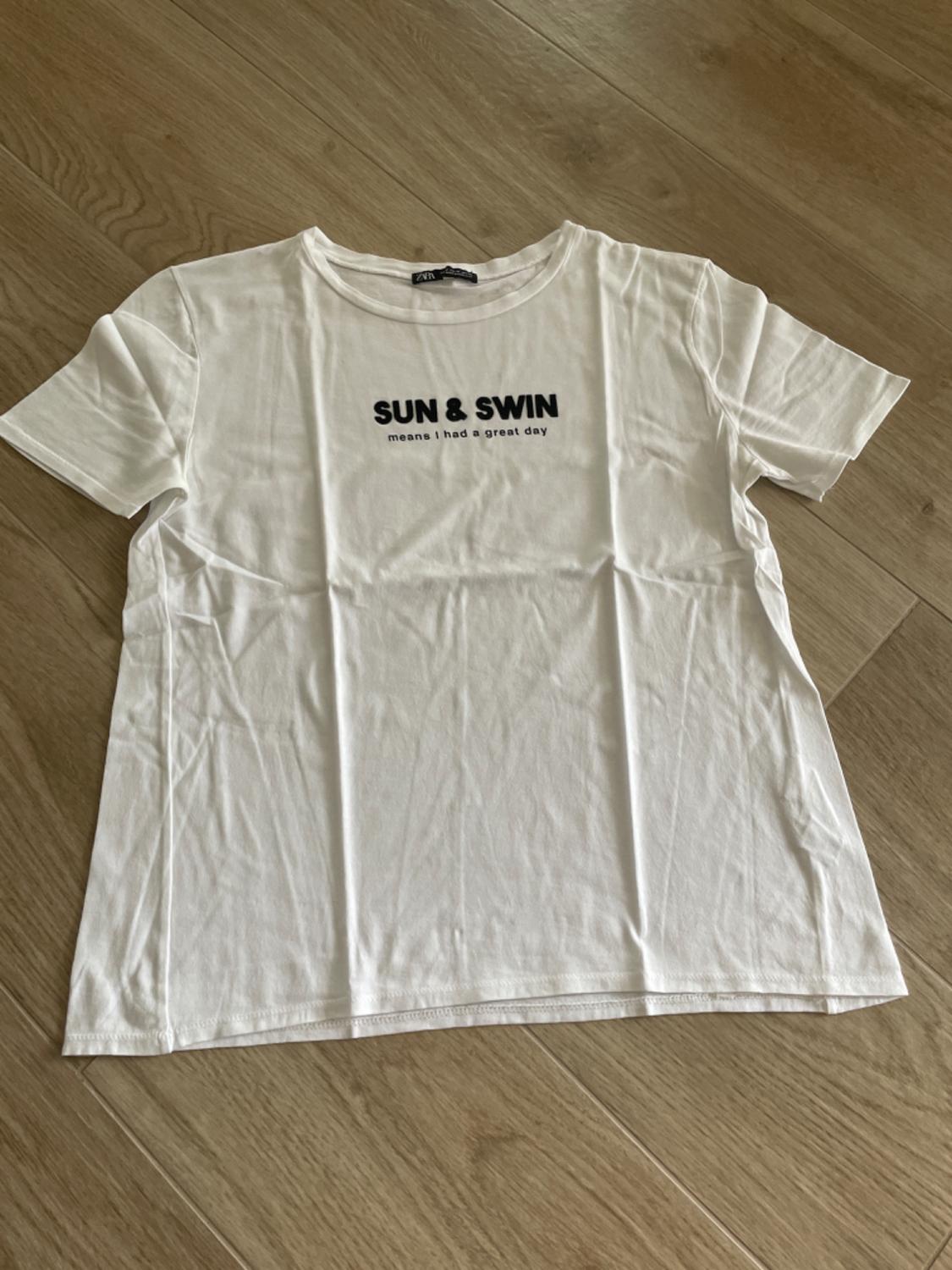 White t-shirt on wooden floor with black text: "SUN & SWIM means I had a great day".