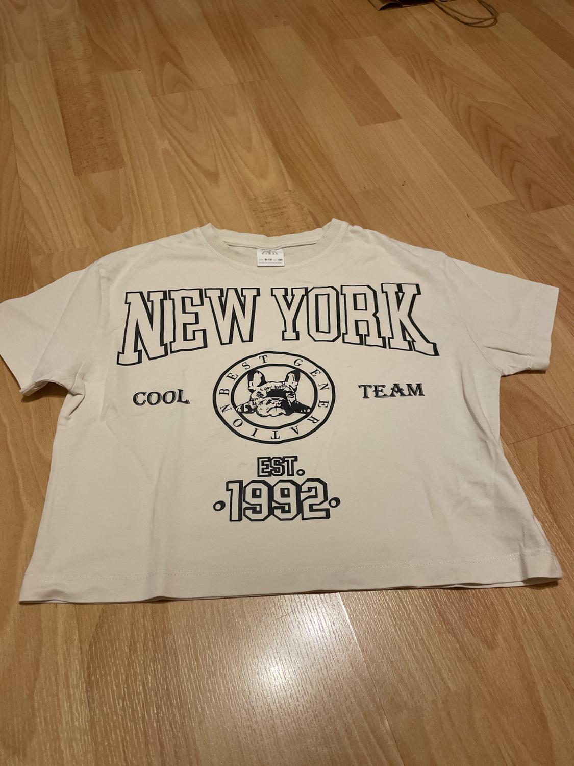 White T-shirt with text "NEW YORK", "COOL TEAM", "EST. 1992" and a bulldog graphic in the centre, on a light wooden floor.