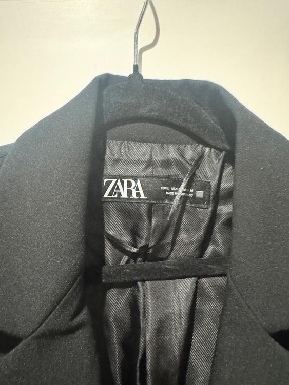 Black jacket hanging on a hanger, with a visible label that says "ZARA". Light background and soft light on the fabric.