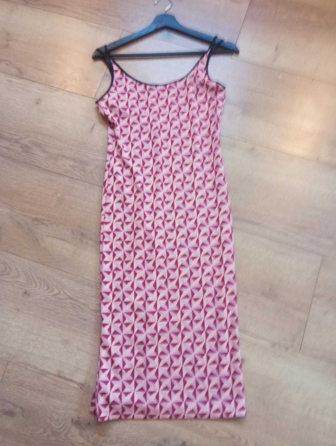 Long black strappy dress with a pink and maroon geometric print, hanging on a black hanger over a wooden floor.