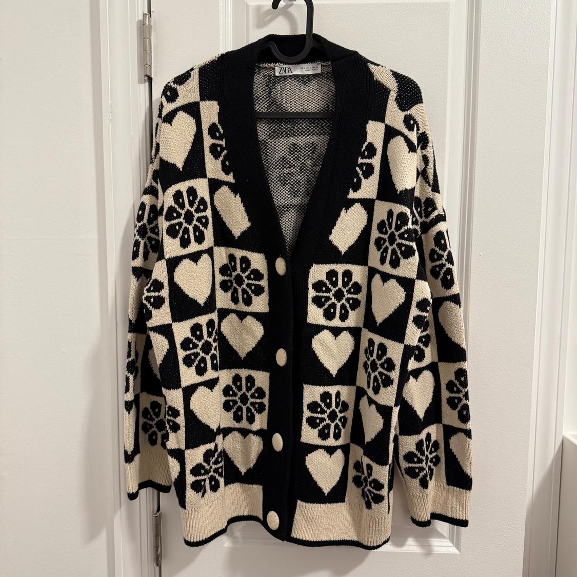 Cardigan hanging on a door, with a black and white checkered design, decorated with hearts and flowers, and large buttons.