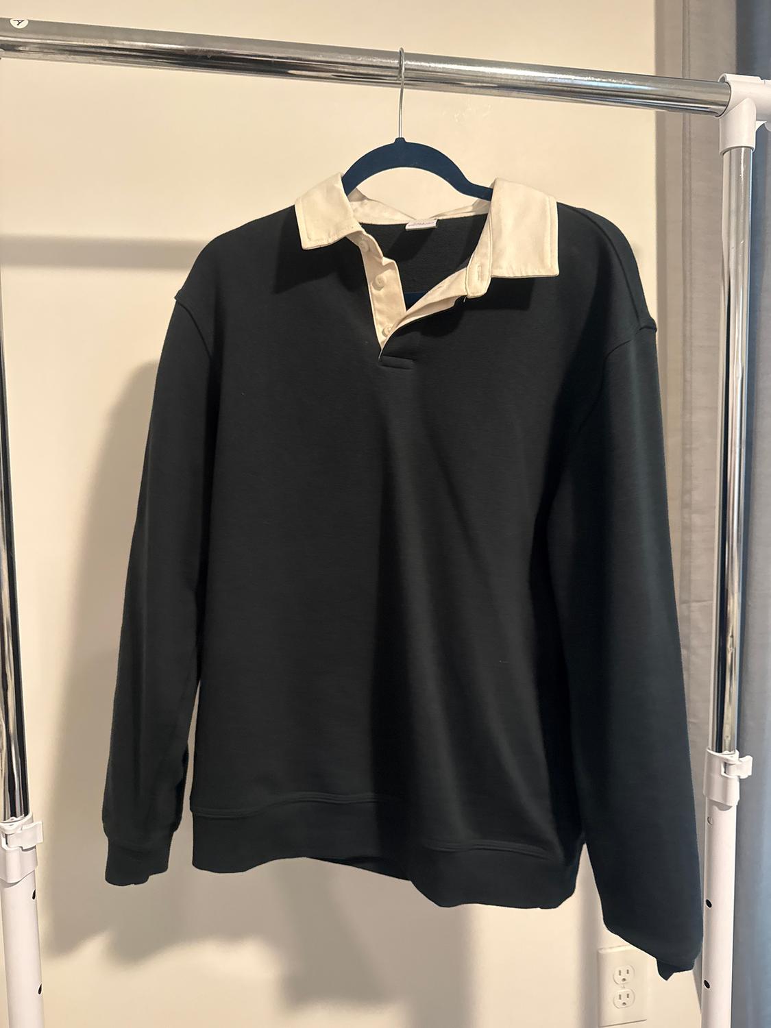 Black sweatshirt with white collar hanging on a blue hanger on a metal stand in a lit room.
