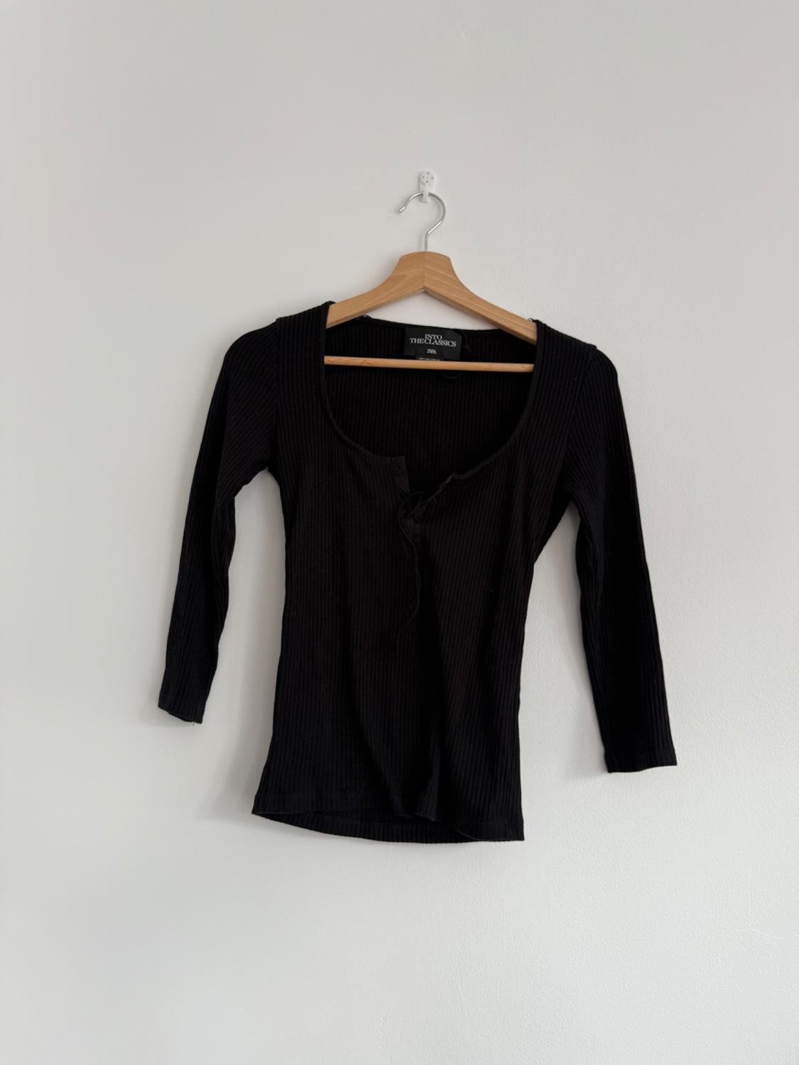 Long-sleeved black jumper hanging on a wooden hanger against a white wall. Label visible: "Two The Classics".