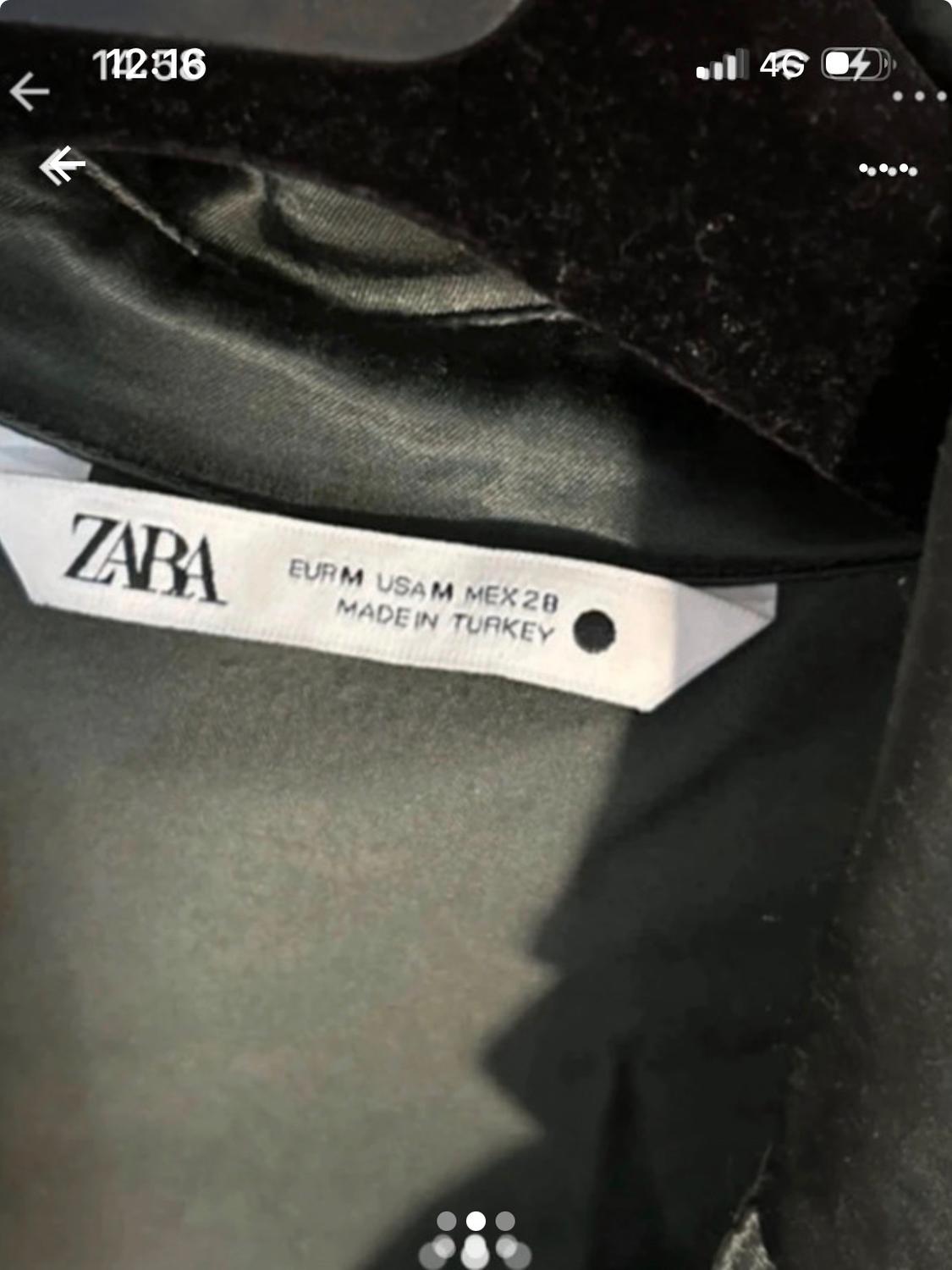 Garment label with the text "ZARA EUR M USA M MEX 28 MADE IN TURKEY" on dark fabric. Phone screen visible.