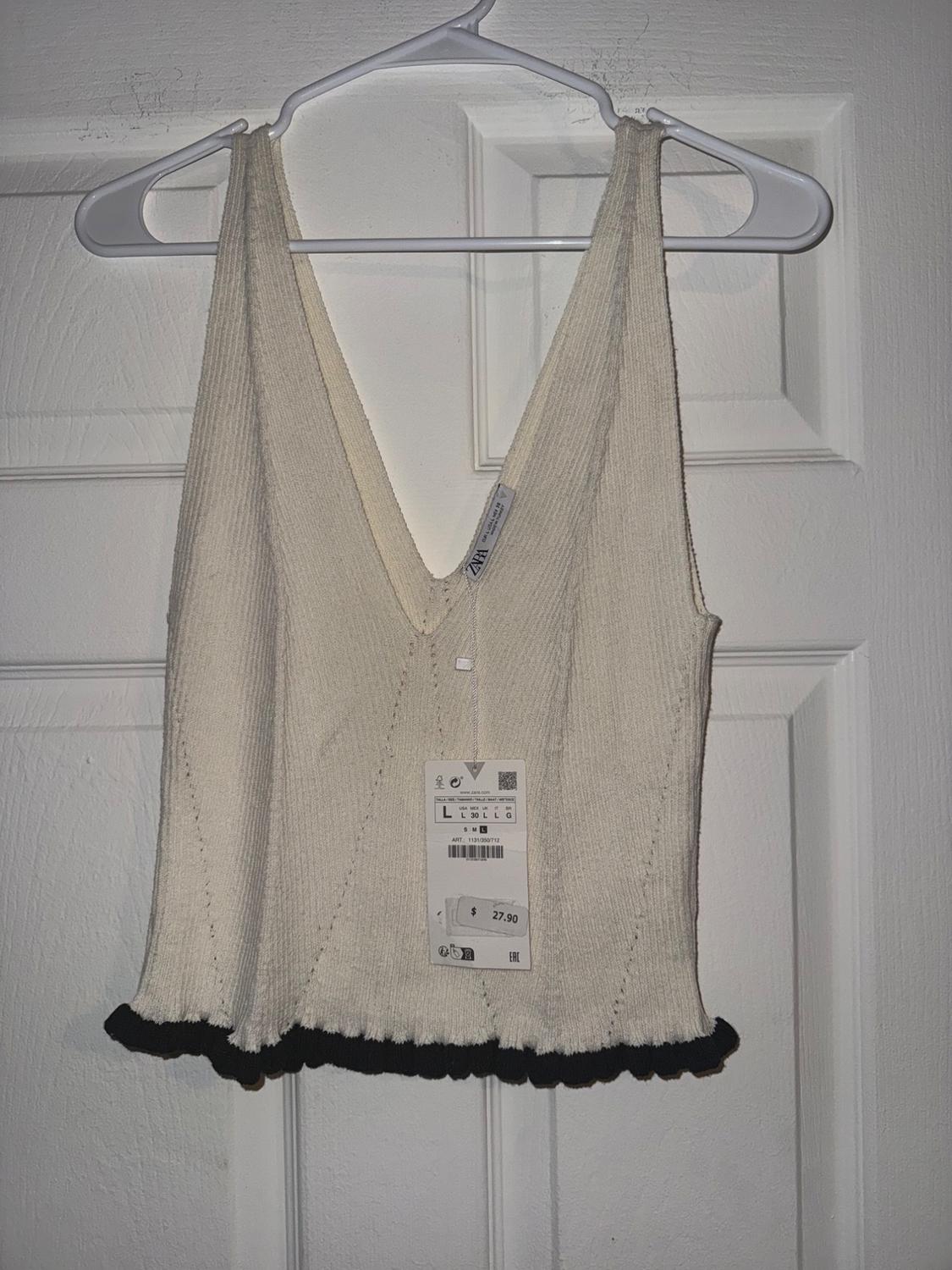 Beige knitted V-neck top with black trim, hanging on a white hanger. Visible tag with price of $27.90.
