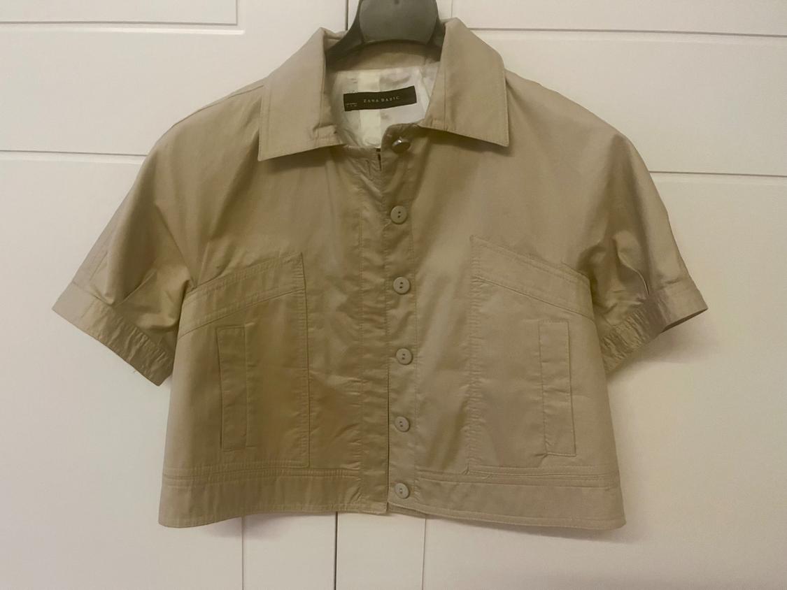 Short-sleeved beige shirt with front buttons and two large pockets, hanging on a hanger in front of a white door.