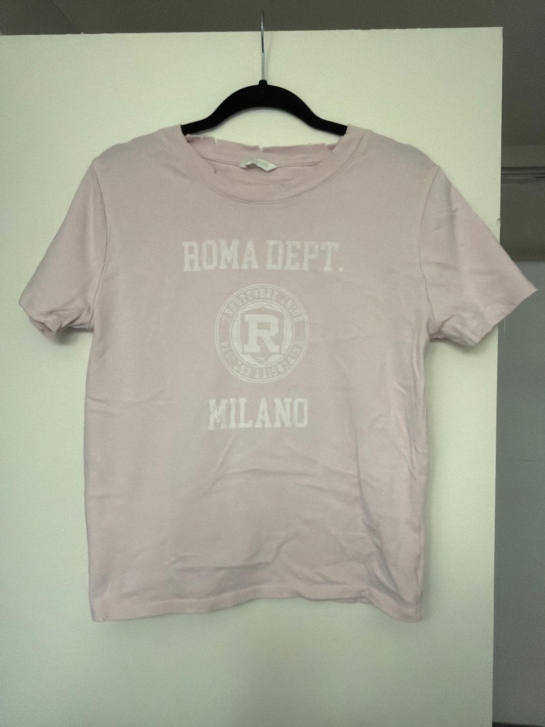Light pink t-shirt hanging on a black hanger, with "ROMA DEPT. MILANO" text on the front. White background.