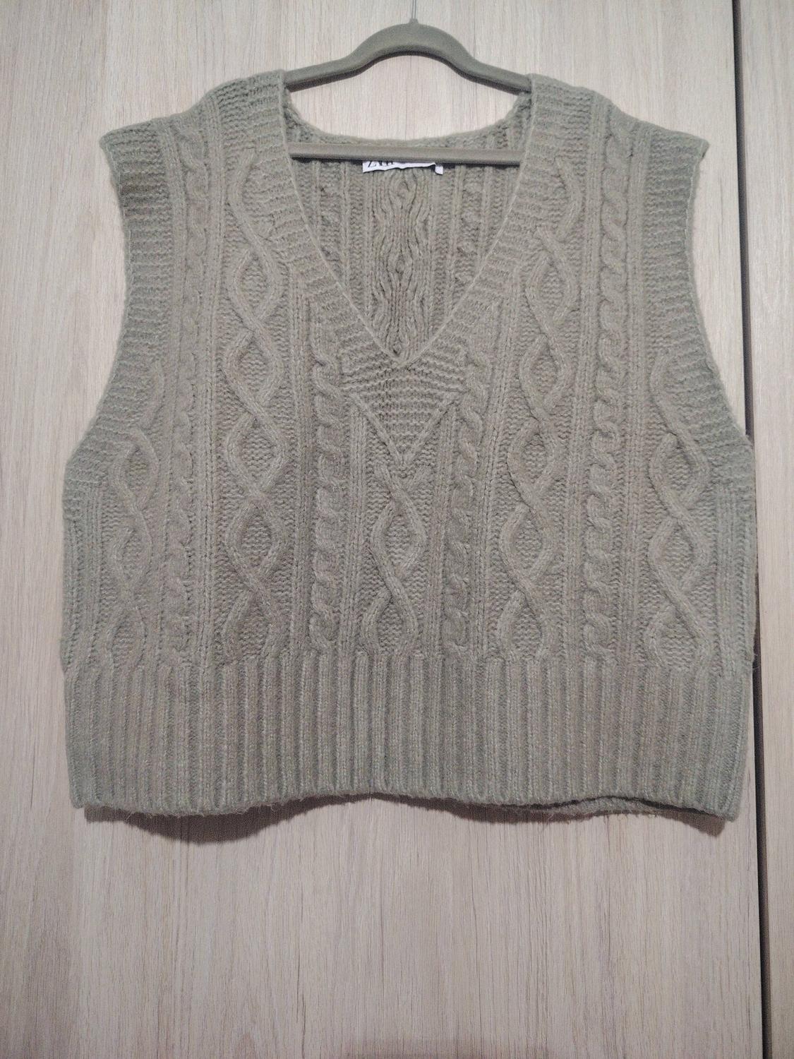 Grey knitted cable-knit waistcoat hanging on a hanger against a light wooden background.