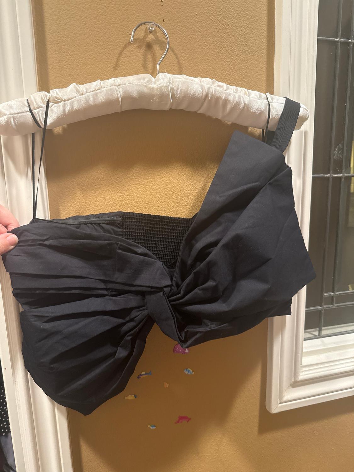 Black top with a large bow on the front, hanging on a white padded hanger. Beige wall background with small fish figures.