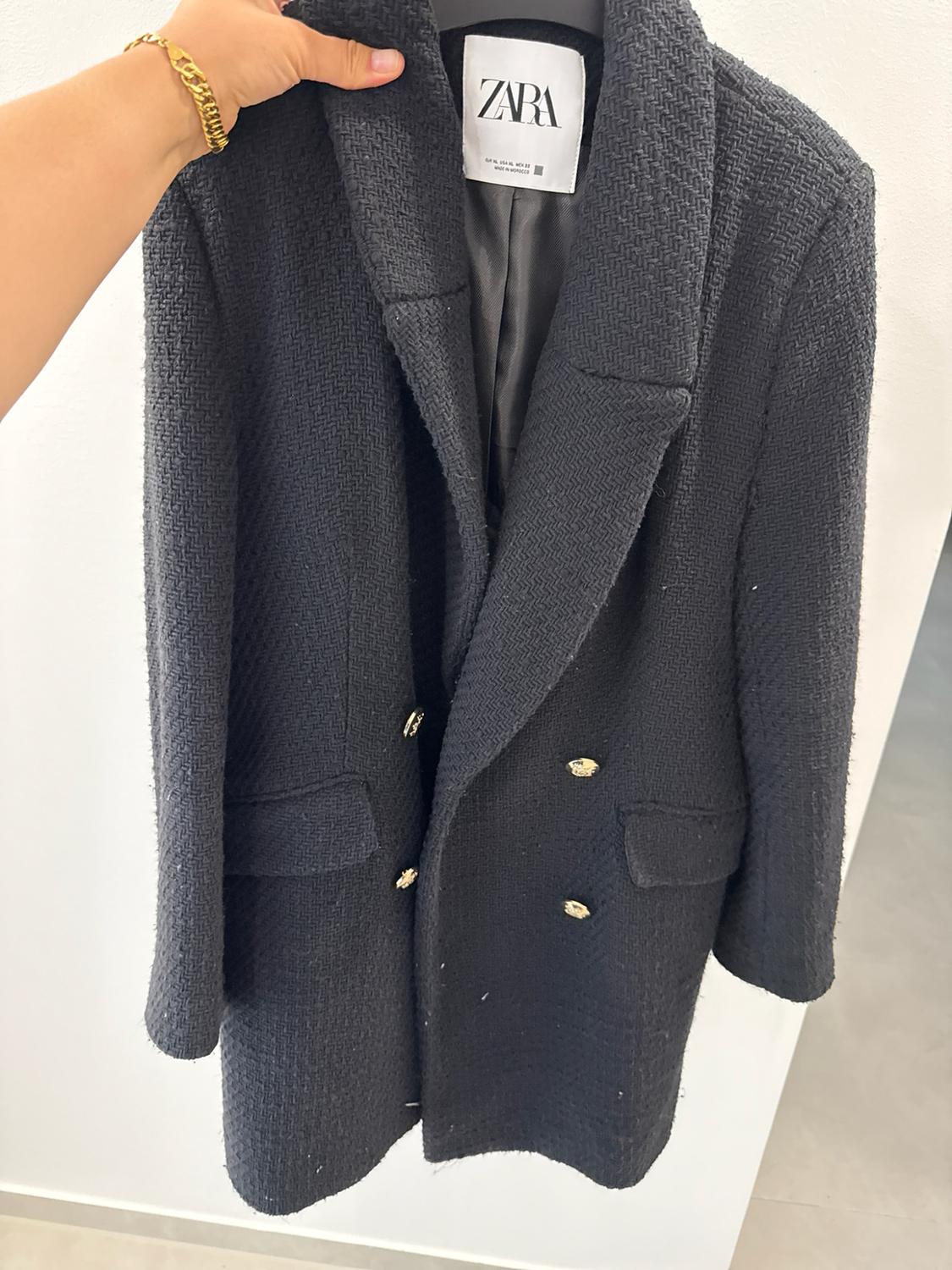 Hand holding a textured black coat with gold buttons. Interior label with ZARA text. White background.
