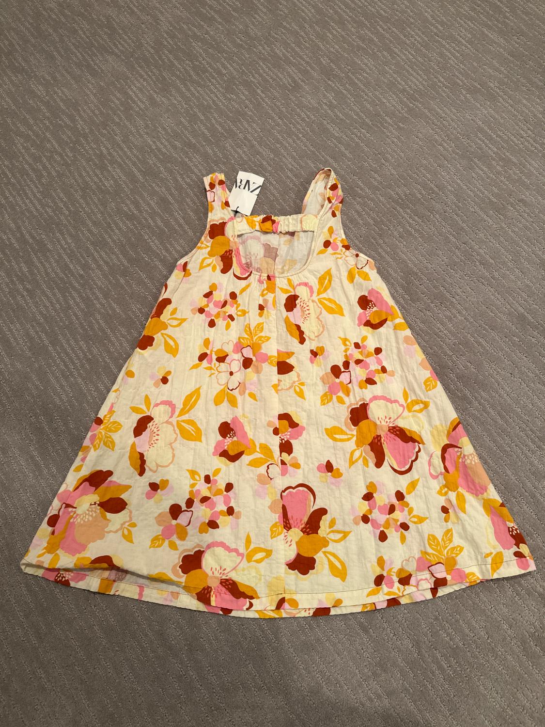 Sleeveless children's dress with a floral print in pink, yellow, and brown tones on a beige background, on a gray surface.