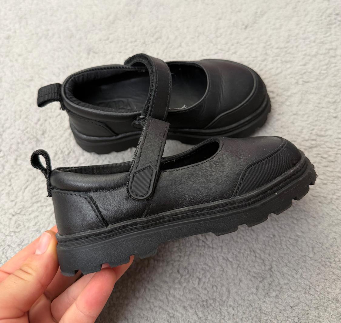 Hand holding a black shoe with a chunky sole and a strap. The other shoe rests on a textured grey background. Text: ZARA.