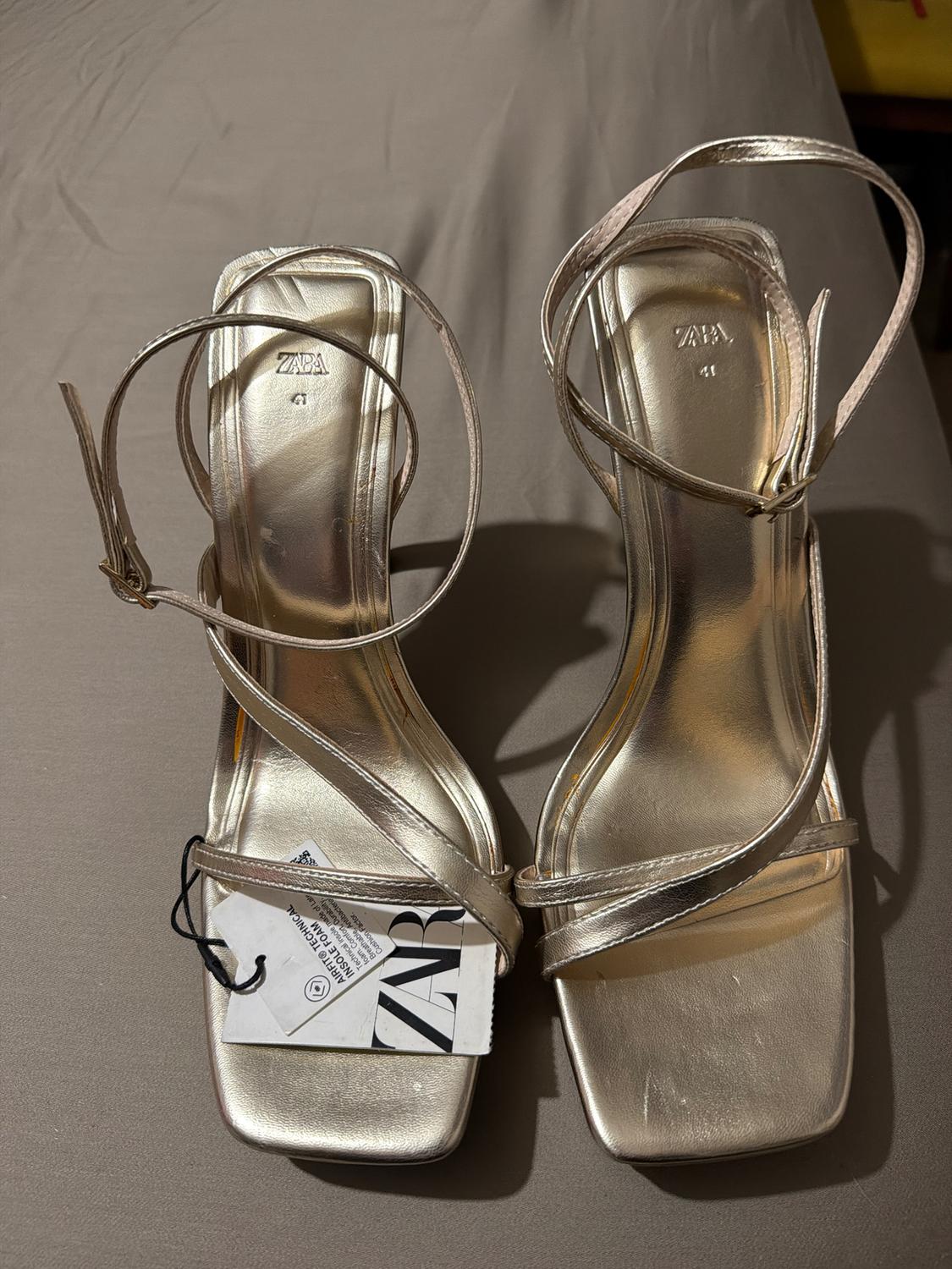 Gold square-heeled sandals with thin straps and a ZARA tag on a beige surface.