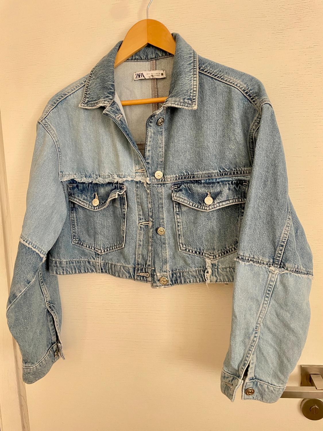 Light blue denim cropped jacket hanging on a wooden hanger, with front pockets and metallic buttons.