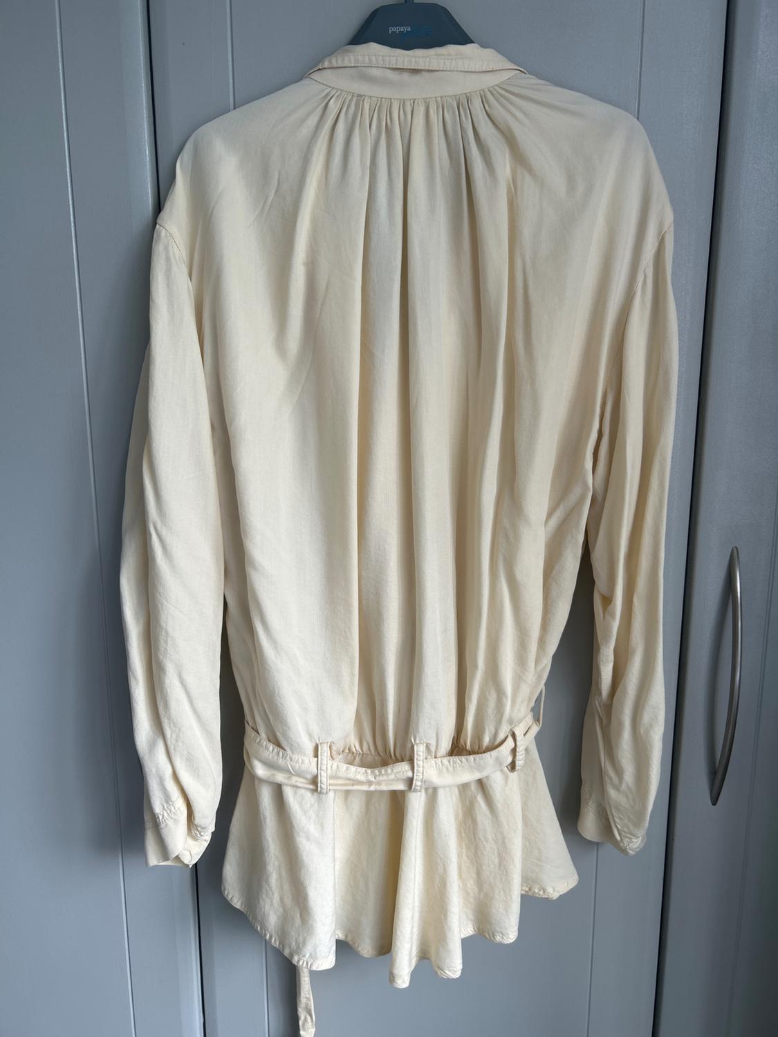 Back view of a cream, long-sleeved, flowing blouse with a belt, hanging in a grey wardrobe. The hanger has the text papaya on it.