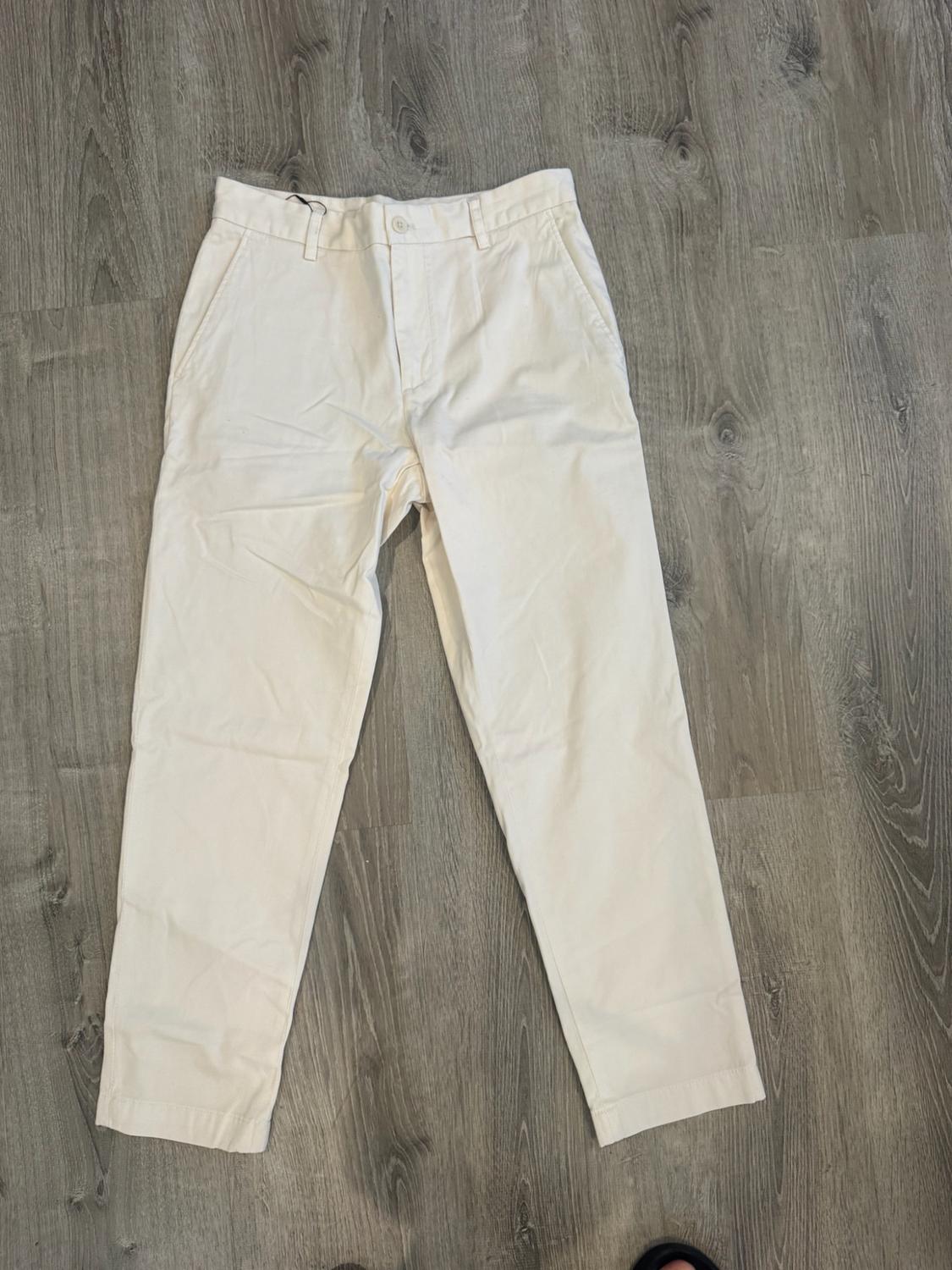Casual ecru trousers with pockets and belt loops, laid out on a grey wooden floor.