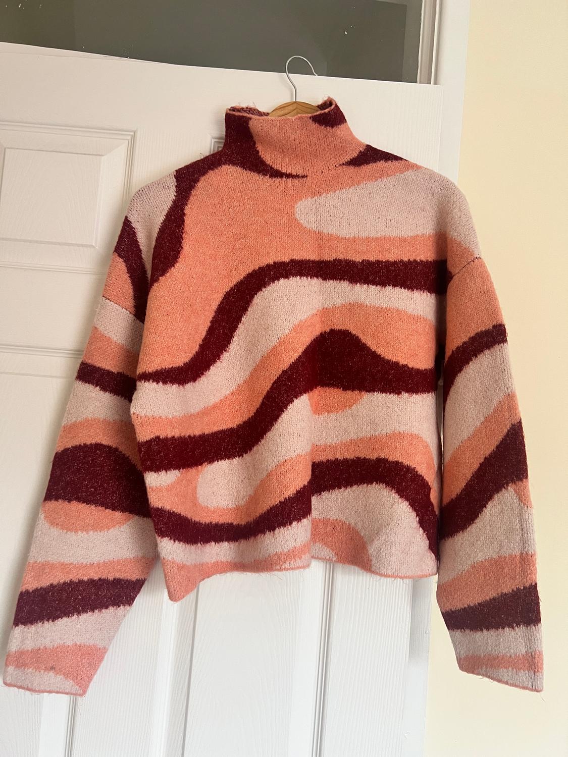 Pink, orange, and burgundy wave-patterned roll-neck jumper, hanging on a white door.