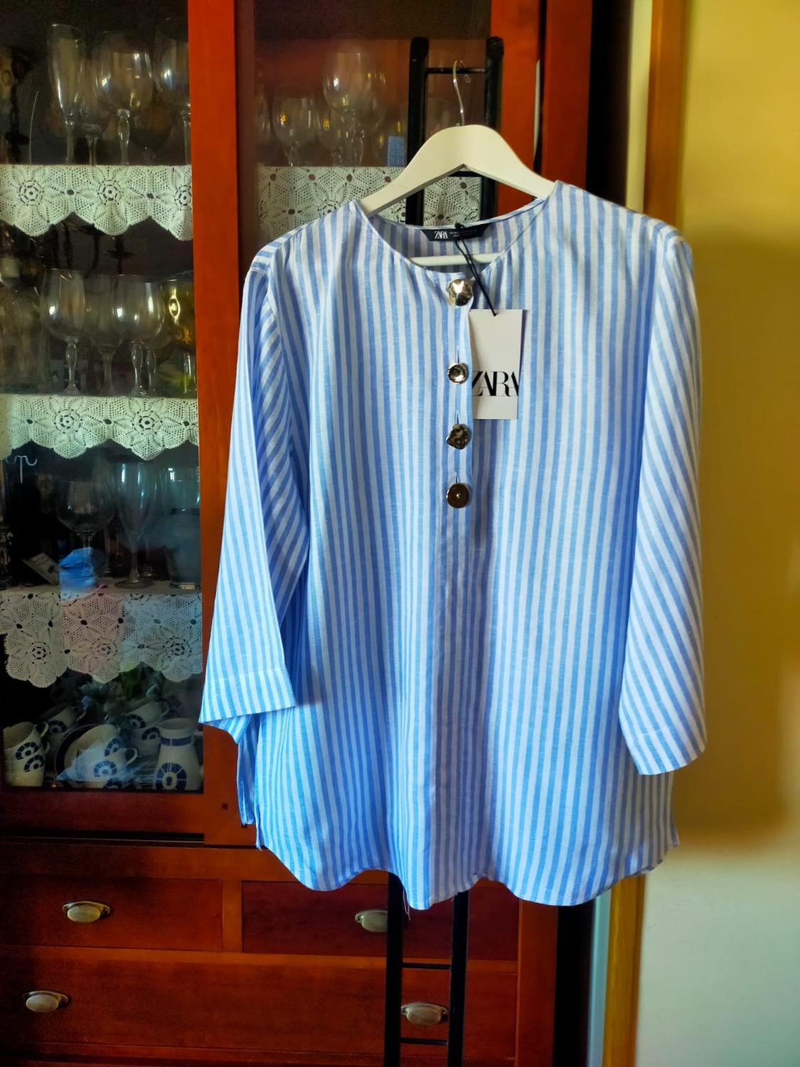 Blue and white striped shirt with large buttons, hanging on a white hanger in front of a wooden cabinet with crystal glasses.