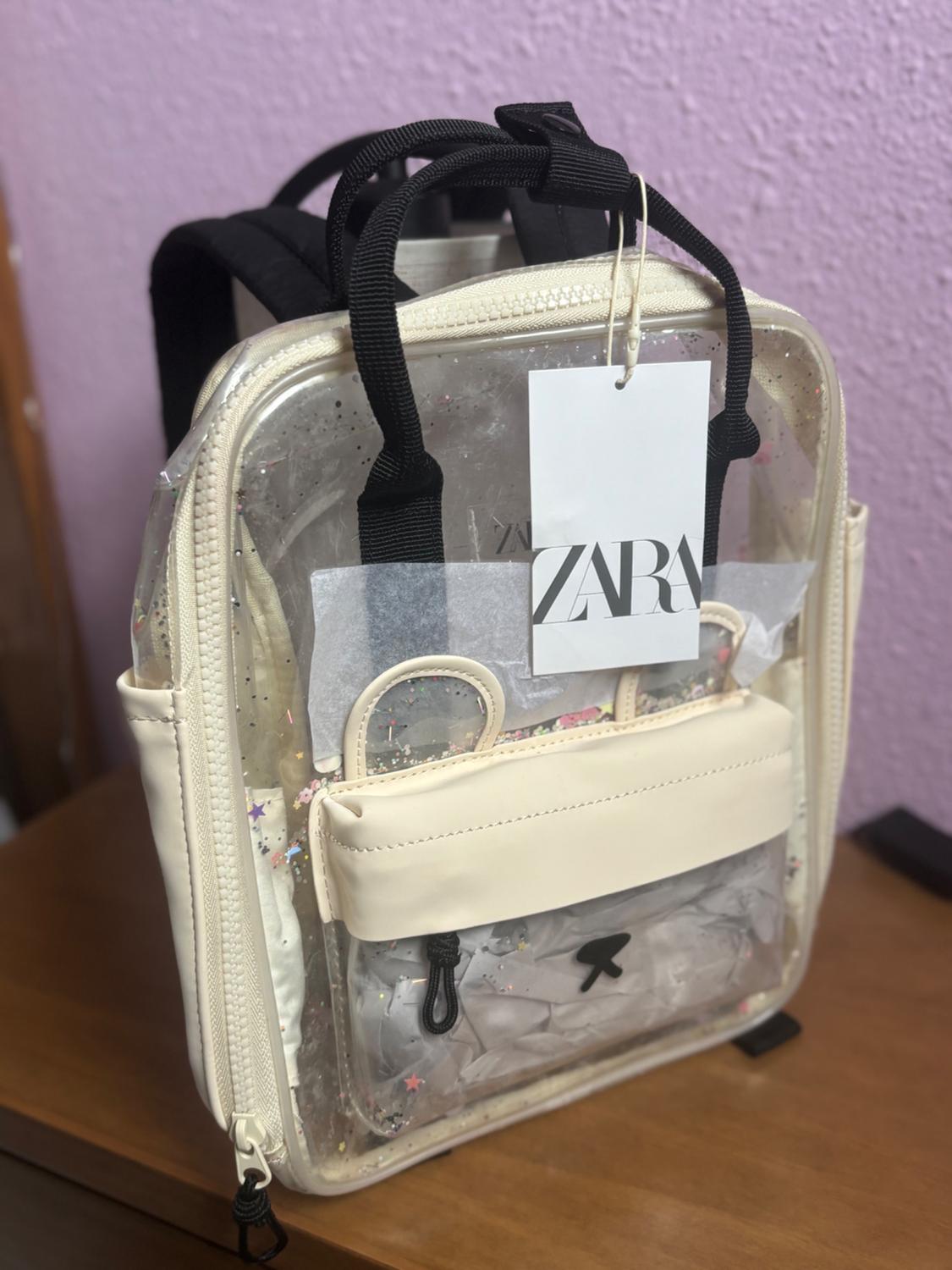 Transparent backpack with confetti details and a beige front pocket. Hanging tag with the text "ZARA". Lilac wall background.