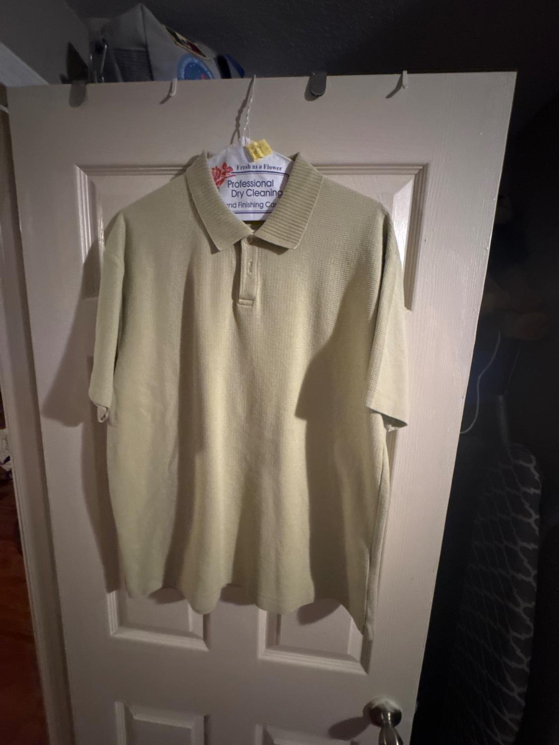 Beige polo shirt hanging on a door with a dry cleaner's hanger. Dark background with a bag at the top.