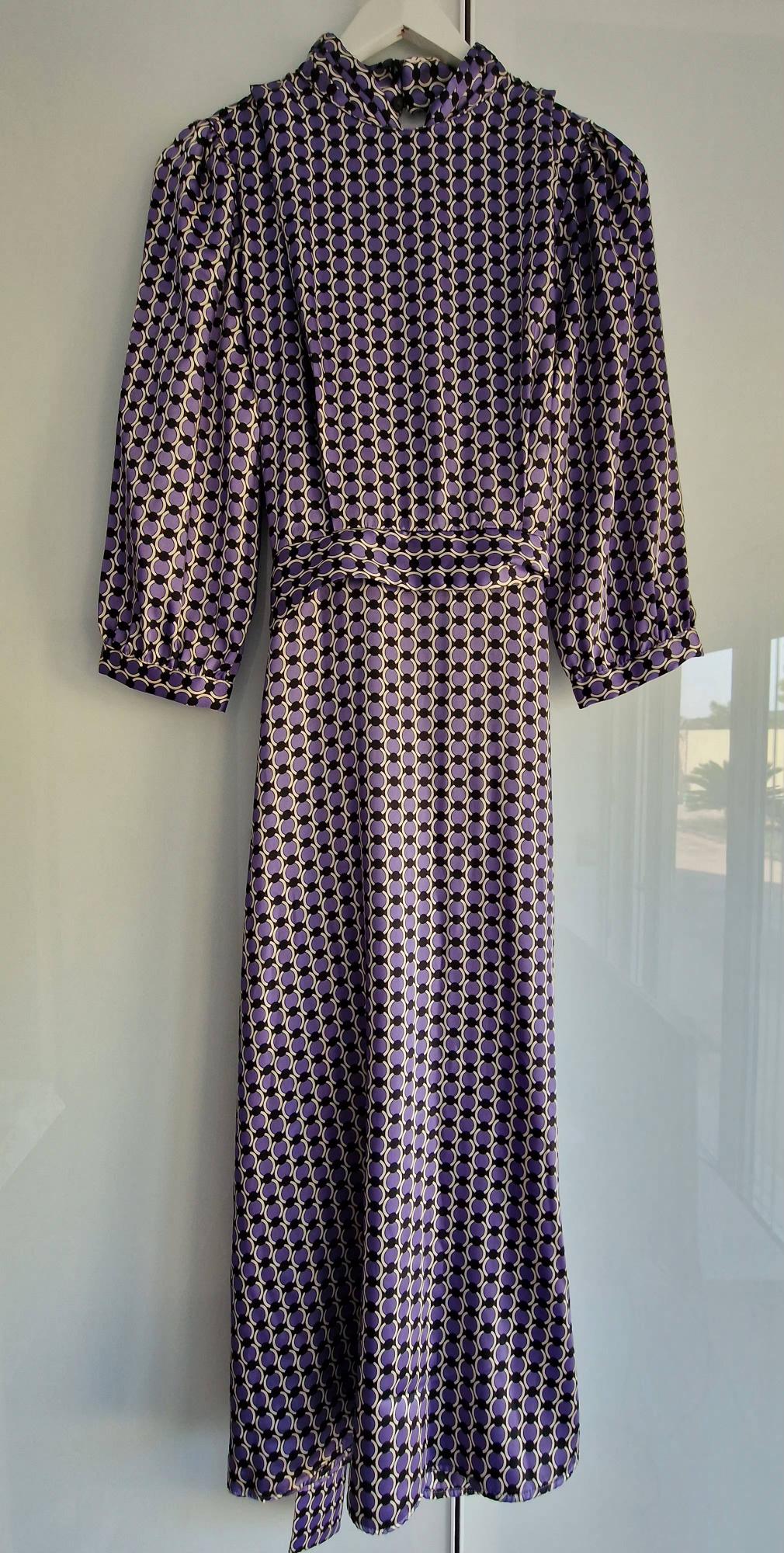Long-sleeved maxi dress with a geometric pattern in purple, black and white, hanging on a white hanger over a glass surface.