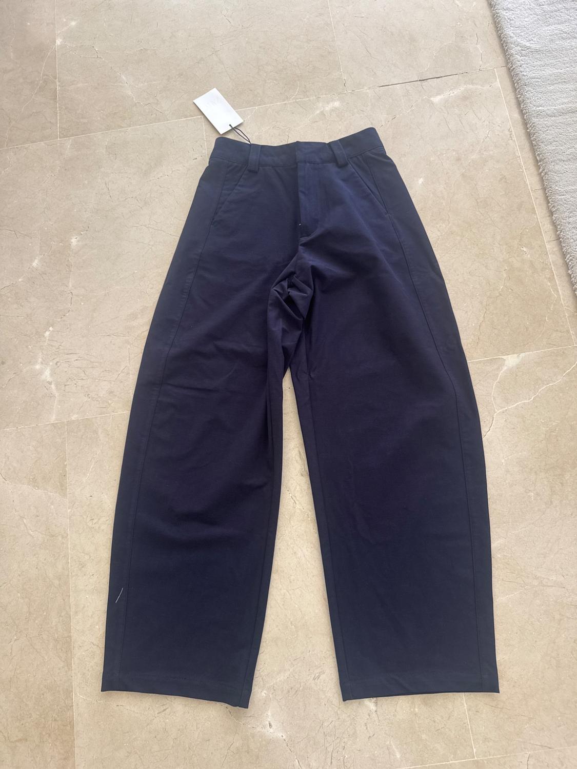 Navy wide-leg trousers with a label, laid out on a light tiled floor next to a rug. Relaxed composition.