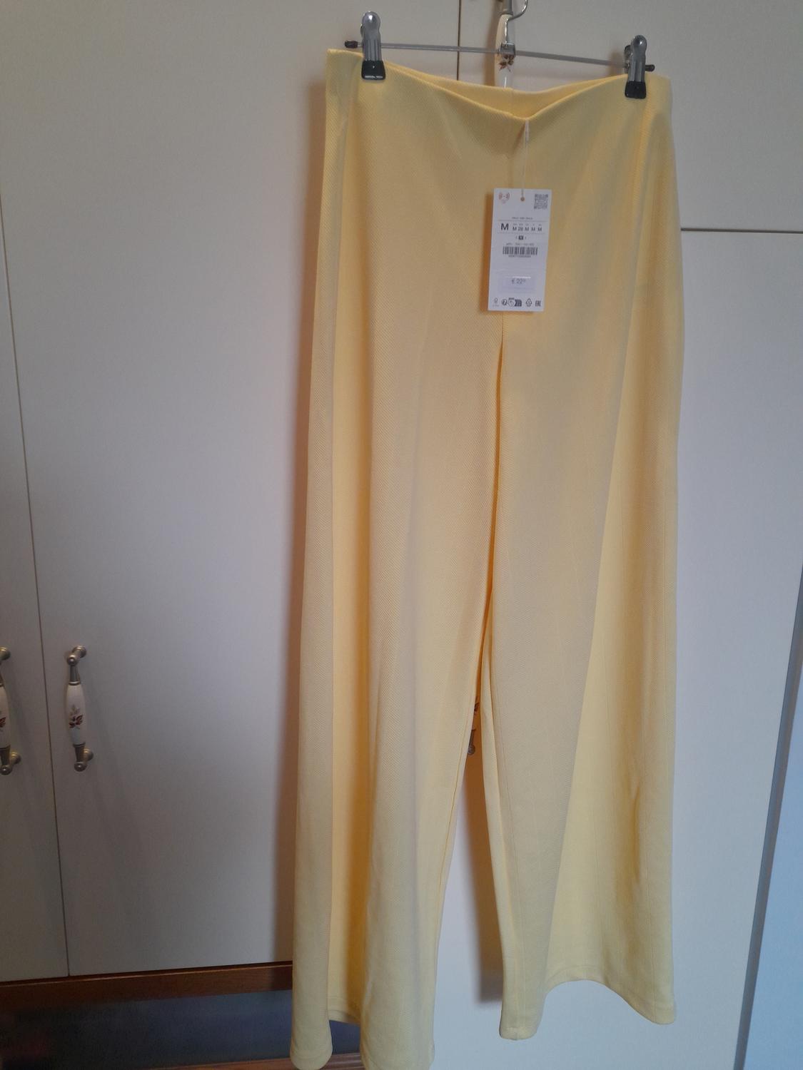 Yellow trousers hanging on a hanger with a visible tag, against a white wardrobe background.