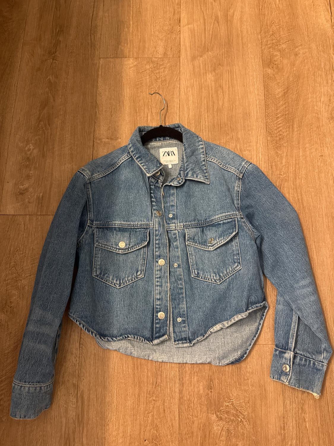 Blue denim jacket with metallic buttons and front pockets, hanging on a hanger over a wooden floor. Visible label: ZARA.