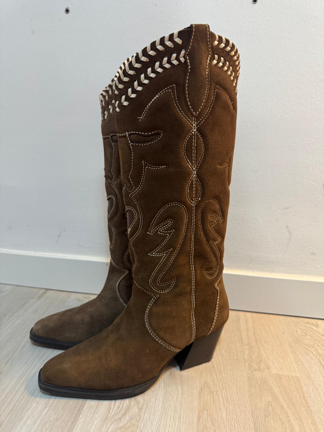 Brown suede knee-high boots with white embroidery and block heel, on a light wooden floor and white background.