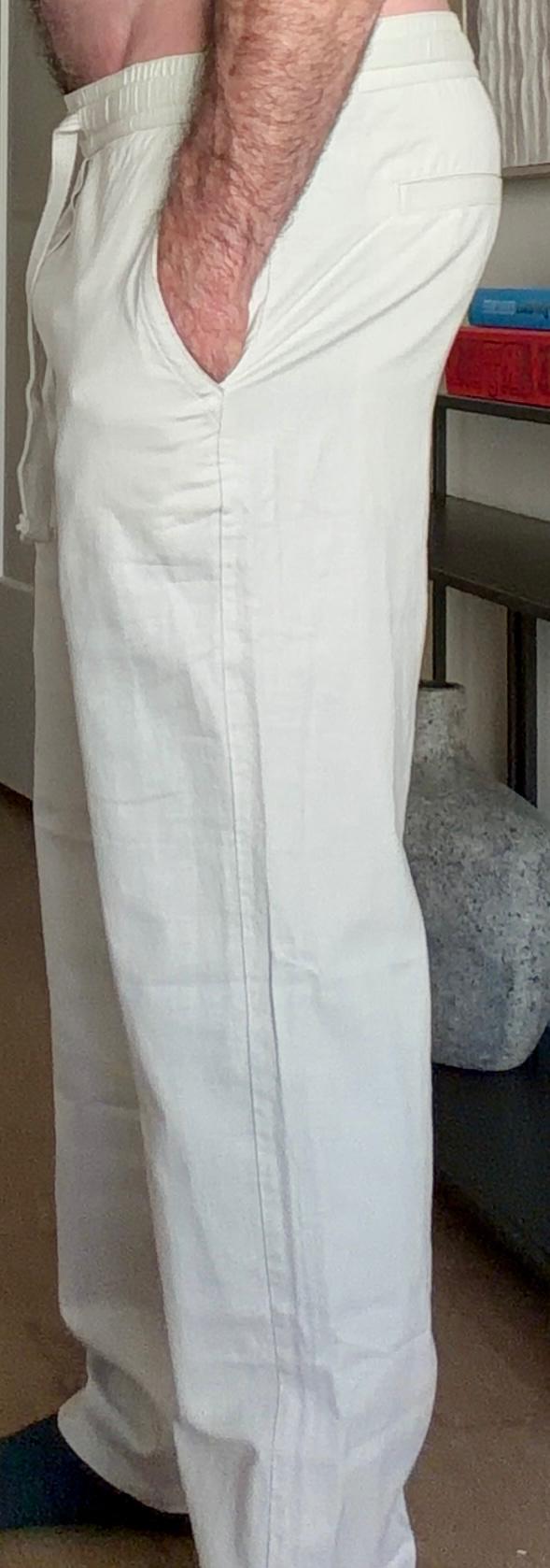 A man is wearing loose white pants with an elastic waistband and has his hand in his pocket. The background is an interior space showing a bookshelf and a vase.