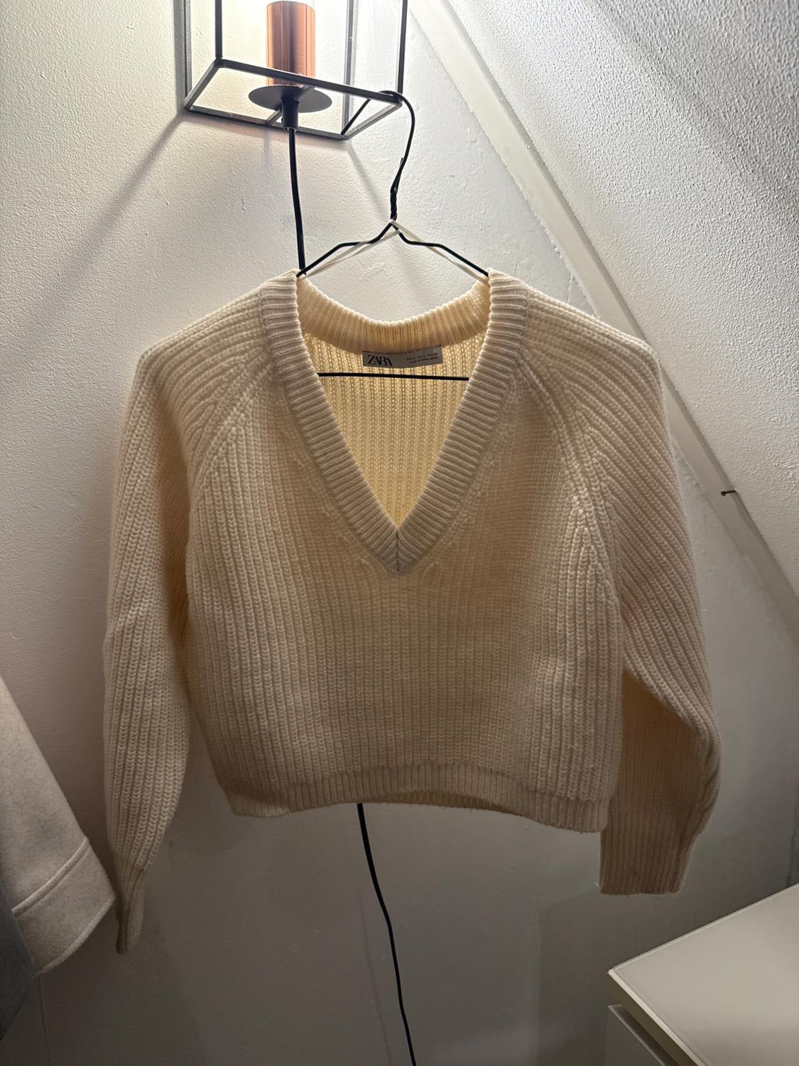 Cream knitted V-neck jumper hanging on a hanger under a wall lamp. Visible label: Zara. Minimalist setting.