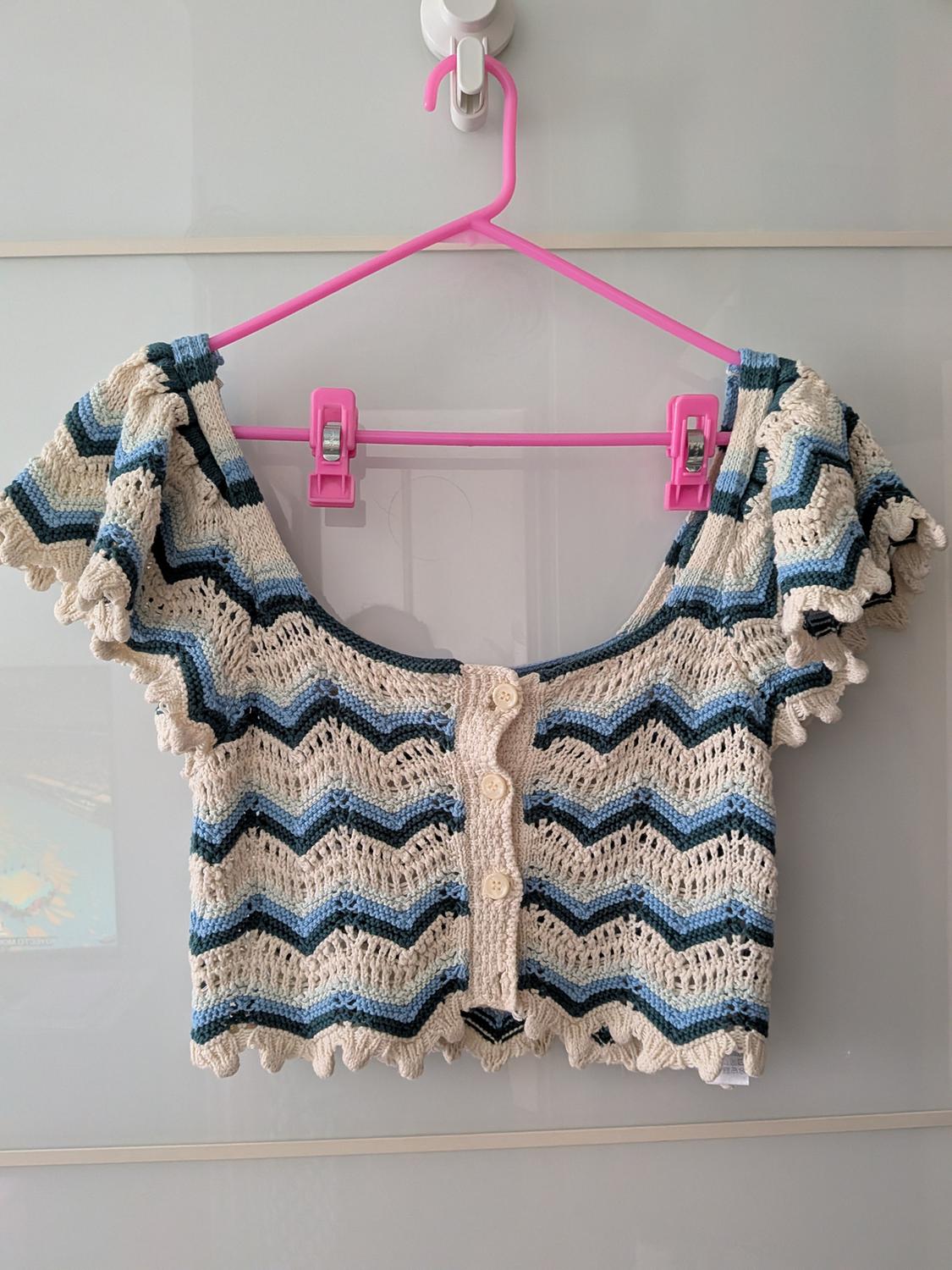 Cream and blue zigzag pattern knit crop top with front buttons and wavy edges. Hanging from a pink hanger against a light background.