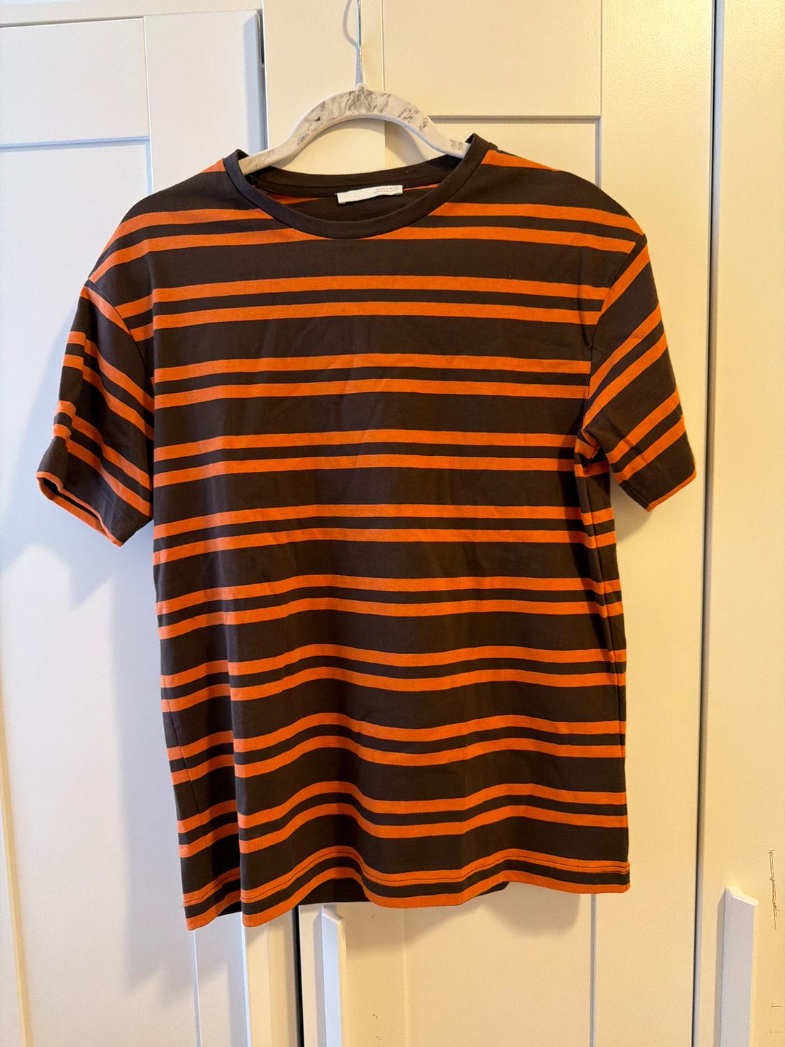 Casual short-sleeved T-shirt with orange and brown horizontal stripes, hanging on a hanger in front of a white wardrobe.