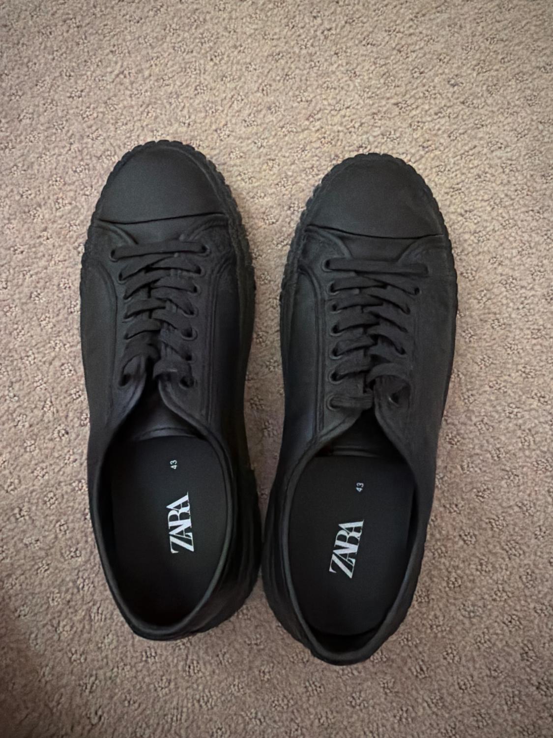 Top-down view of a pair of black sneakers on a beige rug. The insoles read the brand ZARA and size 43.