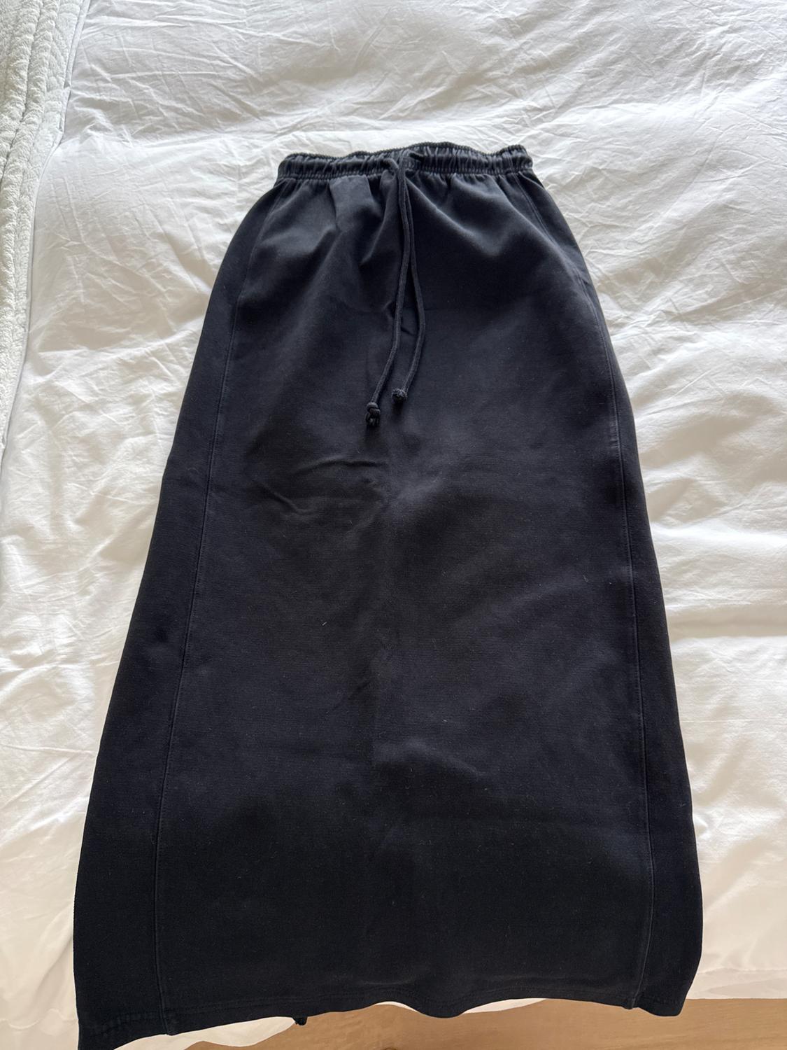Long black skirt with elastic waistband and adjustable drawstring, placed on a bed with wrinkled white sheets.