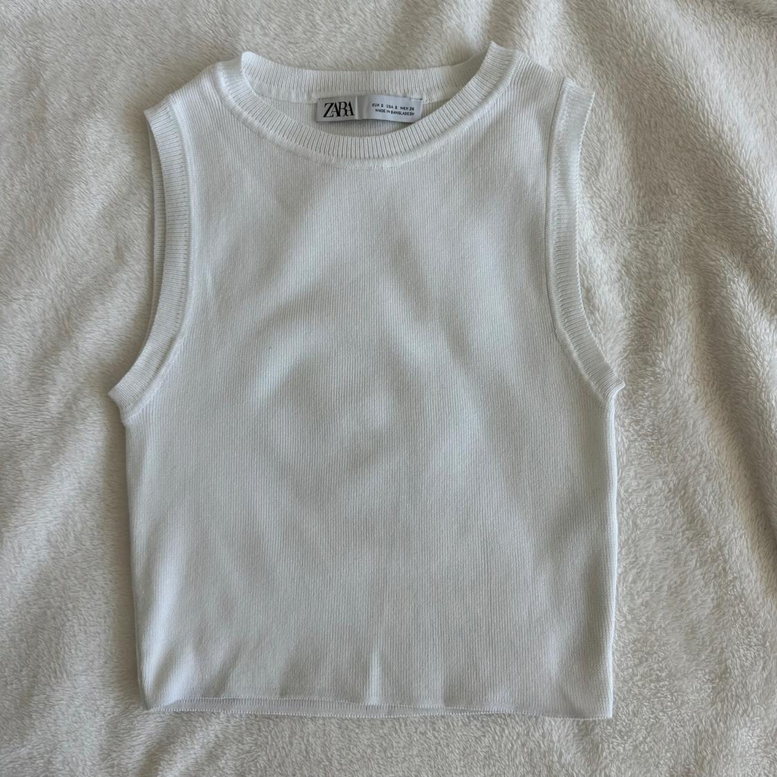 White ribbed knit tank top with visible Zara label, on a soft textured beige fabric surface.