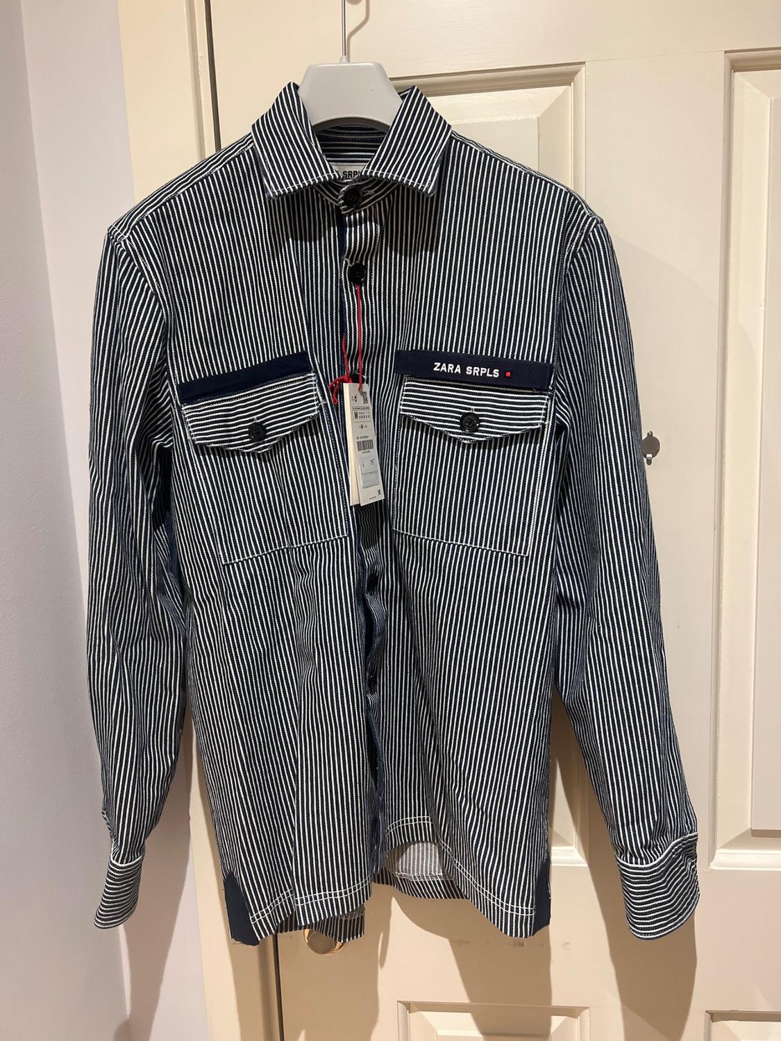 Front view of a blue and white striped shirt with front pockets and text 'ZARA SRPLS'.