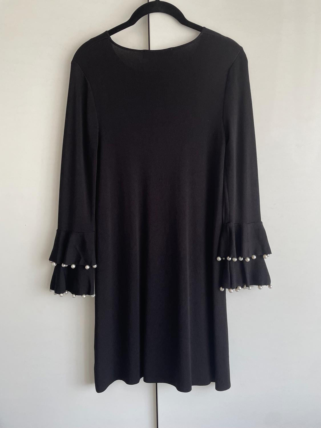 Black long-sleeved dress with ruffles and pearls on the sleeves, hanging on a hanger against a white background.