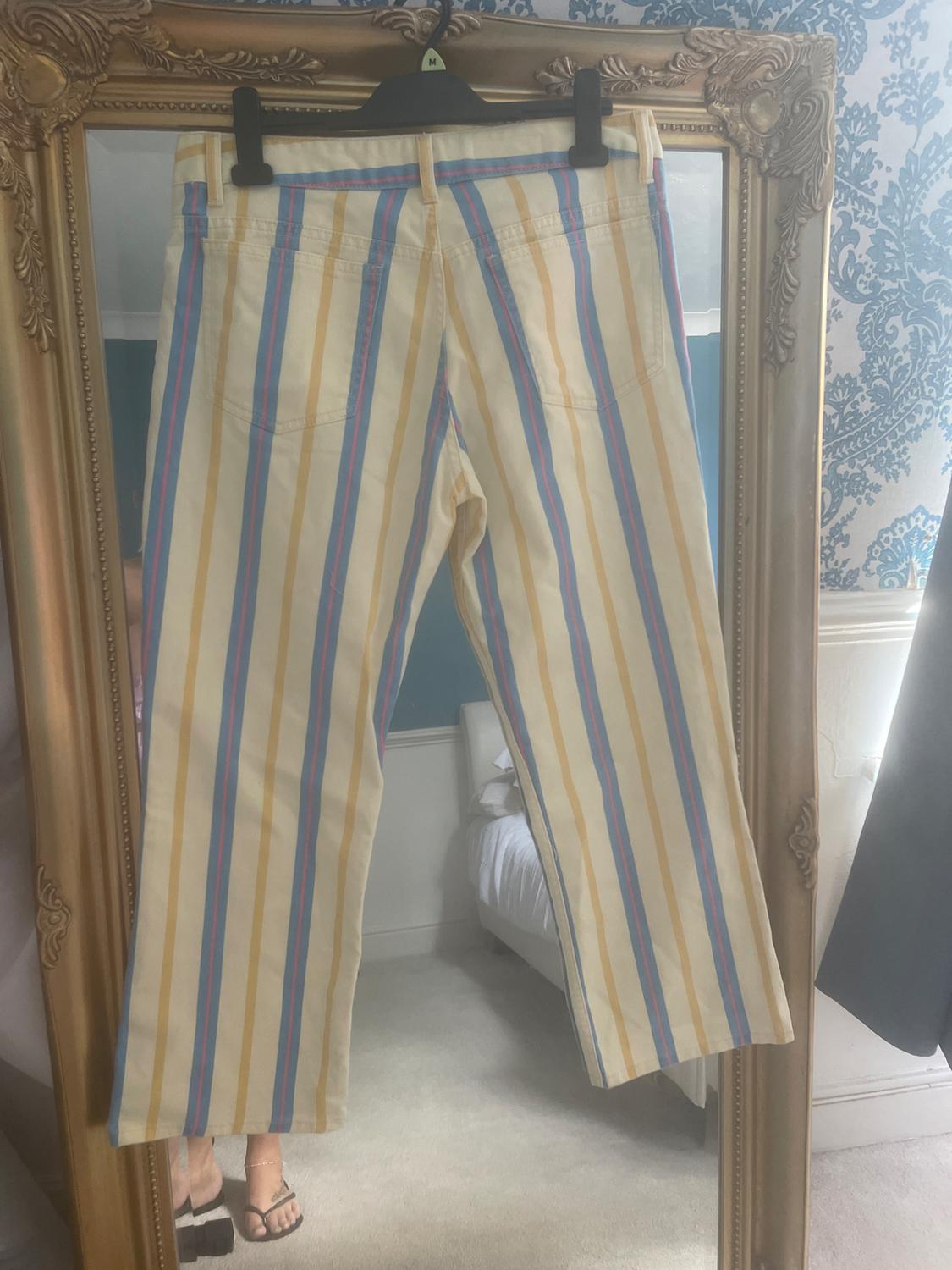 Light-coloured trousers with vertical blue and yellow stripes on a hanger with the letter M, in front of a gold mirror reflecting a bedroom.