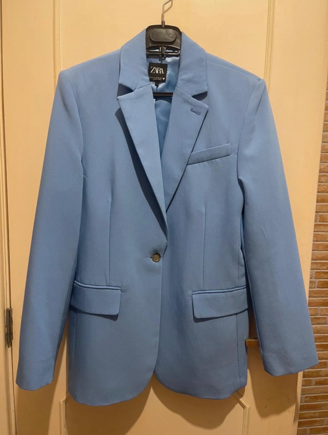 Light blue jacket hanging on a black hanger with a "ZARA" label, over a beige door.